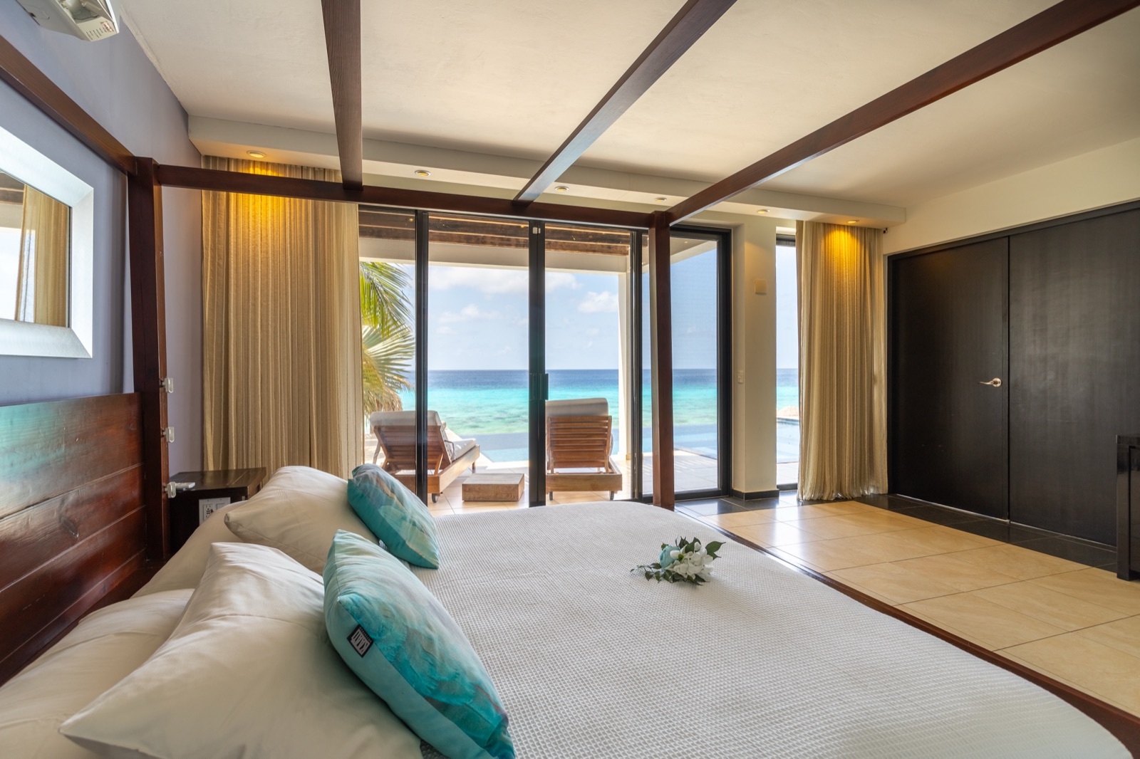 Luxury Bonaire villa master suite with a dark wood canopy bed, floor-to-ceiling glass doors, and a private terrace with lounge chairs overlooking the turquoise Caribbean ocean.